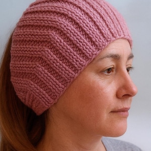 Könnte beinhalten: Ein gestrickter, gerippter Beanie in einem staubigen Rosaton. Der Hut ist aus weichem Garn gefertigt und hat ein eng anliegendes Design. Der Beanie wird auf dem Kopf getragen, wobei das Haar hinten sichtbar ist.