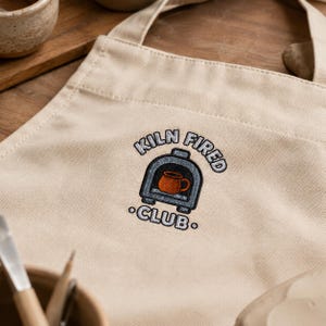 Peut inclure: Un tablier en toile beige avec une broderie représentant un four gris contenant une tasse orange. Le texte KILN FIRED CLUB est cousu en fil noir en arc de cercle autour de l'illustration du four, avec le mot CLUB placé en dessous.