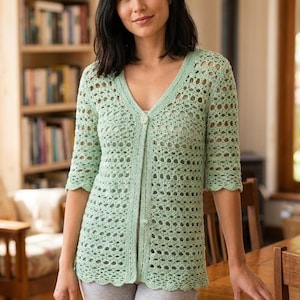 May include: A pale green, open-weave crochet cardigan with a V-neck and three-quarter sleeves. The cardigan has a scalloped hem and is buttoned up the front. The person is wearing grey shorts. The background includes a wooden table and a bookshelf.