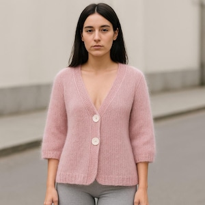 May include: A light pink cardigan with a V-neckline and three-quarter sleeves. The cardigan has two white buttons down the front and a textured knit design. The soft, fuzzy texture of the cardigan is visible.