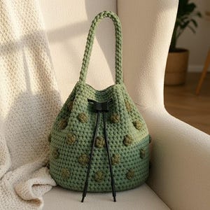 May include: A green crochet bucket bag with a braided handle and drawstring closure. The bag features textured, raised floral-like details and is displayed on a cream-colored chair. The bag's color is a muted, earthy green.