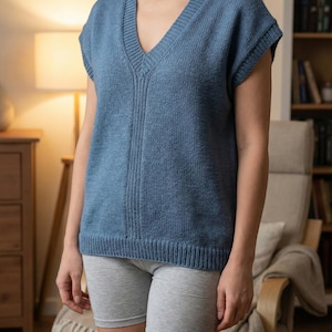 May include: A blue knitted vest with a V-neck. The sleeveless top has a ribbed hem and a vertical ribbed design down the center. The model is wearing gray shorts. The background includes a chair, a lamp, and a bookshelf.