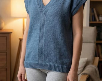 Puede incluir: Un chaleco azul tejido con cuello en V. La parte superior sin mangas tiene un dobladillo acanalado y un diseño acanalado vertical en el centro. La modelo lleva pantalones cortos grises. El fondo incluye una silla, una lámpara y una estantería.