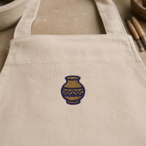 May include: A beige canvas apron featuring a centered embroidered design of a gold-colored vase with a dark blue rim and a decorative dark blue geometric pattern around the middle section of the vessel, showcasing clean stitching and fabric texture.