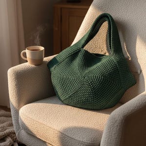 May include: A dark green, hand-crocheted tote bag with a rounded shape and wide straps sits on a cream-colored armchair. A beige and brown mug with a hot beverage rests nearby. The bag's texture is visible, suggesting a handmade item.