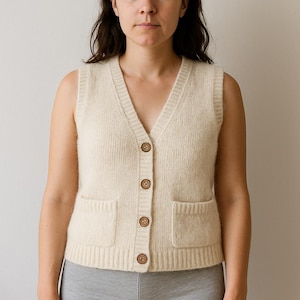 Puede incluir: Chaleco de punto sin mangas de color crema con cuello en V y botones de madera. El chaleco tiene dos bolsillos delanteros y está hecho de un material suave y texturizado. La prenda forma parte de una colección de ropa.