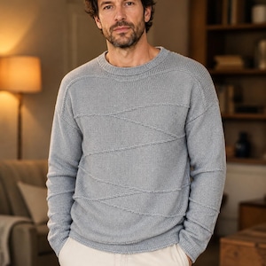 Può includere: Un maglione azzurro con girocollo e maniche lunghe. Il maglione presenta un design a maglia strutturata con accenti cuciti diagonali sul petto. La persona nell'immagine indossa pantaloni bianchi.