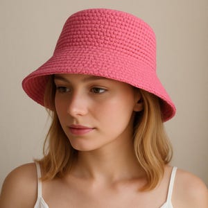 Puede incluir: Un sombrero de pescador rosa de ganchillo. El sombrero tiene una corona redondeada y un ala ligeramente inclinada hacia abajo. El sombrero está hecho de un material texturizado y tejido. El sombrero es de un tono rosa vibrante y parece hecho a mano.