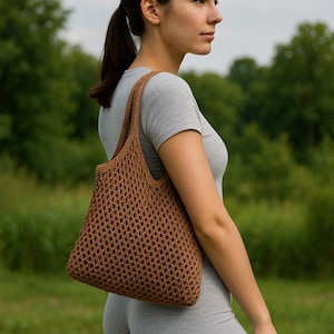 Puede incluir: Un bolso tote marrón de tejido abierto con una correa para el hombro. El bolso está hecho de un material tejido y tiene una gran capacidad. El bolso lo lleva una persona que lleva una camiseta gris y leggings.