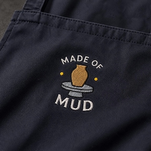 May include: A dark navy blue apron featuring an embroidered design in the center. The embroidery depicts a gold-colored pottery vase on a grey potter's wheel, surrounded by the white text "MADE OF MUD" and two small yellow dots.
