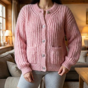 Könnte beinhalten: Ein hellrosa Cardigan-Pullover mit Rippstrickmuster. Der Pullover hat einen runden Ausschnitt, lange Ärmel, zwei Vordertaschen und dekorative Knöpfe. Die Knöpfe sind silberfarben mit kristallartigem Aussehen. Das Model trägt graue Shorts.