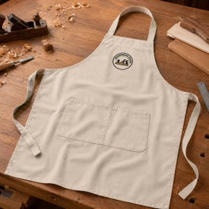 Pode incluir: Um avental bege com alça de pescoço, dois bolsos frontais e um logótipo circular no peito que apresenta o texto WOODWORKING STUDIO juntamente com uma ilustração de uma plaina. O avental tem laços laterais compridos para prender a peça.