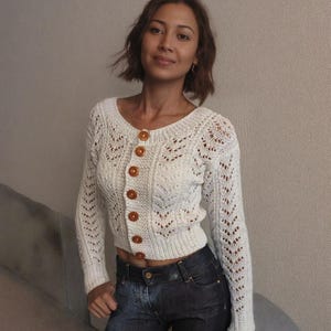 Può includere: Un cardigan bianco lavorato a uncinetto con scollo rotondo e bottoni in legno. Il cardigan presenta un delicato motivo a pizzo ed è corto in vita.