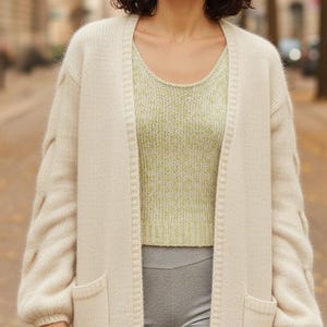 Op de afbeelding: Een vrouw draagt een crèmekleurig lang vest met zakken en een lichtgroene top. Het vest heeft getextureerde mouwen. Ze draagt ook grijze leggings. De achtergrond is een vage buitenscène.