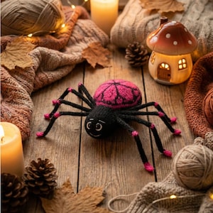 May include: A handmade black and pink spider toy with a smiling face, set on a wooden surface with autumn leaves, pine cones, and a mushroom-shaped candle holder. The spider has a pink and black patterned body and pink accents on its legs.