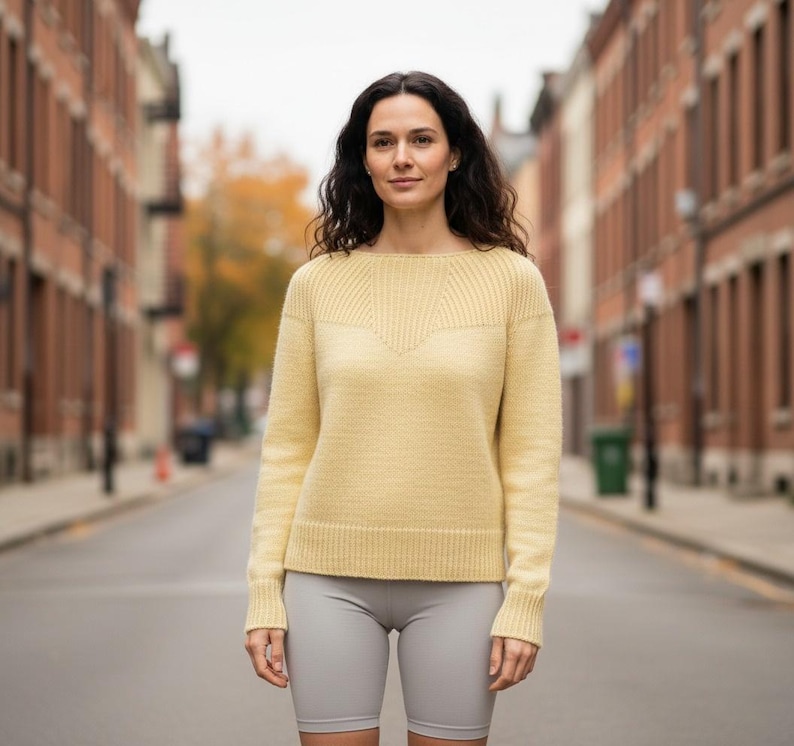 May include: A light yellow sweater with a ribbed design on the upper chest and shoulders. The sweater has long sleeves and is paired with gray shorts. The background shows a city street with brick buildings.