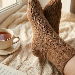 May include: A pair of brown knitted socks with a detailed leaf pattern. The socks are resting on a cream-colored cable-knit blanket, with a cup of tea and a book visible in the background. The socks appear to be made of a soft, warm material.