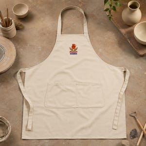 May include: A beige apron featuring a central graphic of two hands holding a small orange clay pot, with the text "MUDDY HANDS" printed in dark blue and red below the illustration. The apron includes a large front pocket with two compartments.