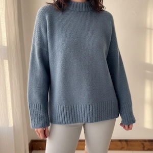 Peut inclure: Un pull bleu clair avec un col montant et des poignets côtelés. Le pull a des manches longues et une coupe légèrement ample. La personne porte un short de cycliste gris clair. Le pull est en maille douce et texturée.