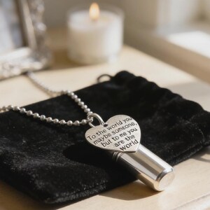 May include: A silver-colored pendant necklace with a heart-shaped charm and a cylindrical vial. The heart charm has the engraved text: "To the world you maybe someone, but to me you are the world." The necklace rests on a black velvet pouch.