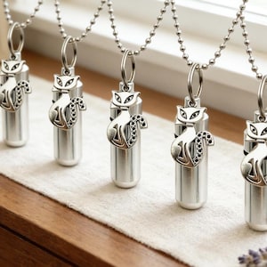 May include: Five silver-tone cat pendant necklaces displayed on a wooden surface. Each necklace features a cylindrical container with a cat charm. The cat charms have detailed features and are attached to a ball chain.