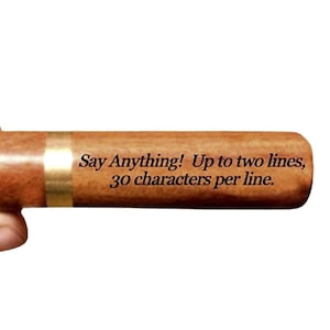 May include: A cylindrical brown wooden object with a smooth finish and a metallic gold band. The surface features black printed text that reads: Say Anything! Up to two lines, 30 characters per line. The object has a rounded end and a uniform shape.