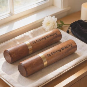 May include: Two wooden urns with brass bands and the inscription "In Loving Memory" are displayed on a white cloth. A lit candle, a black velvet bag, and a white flower are in the background. The urns are cylindrical and have a polished finish.