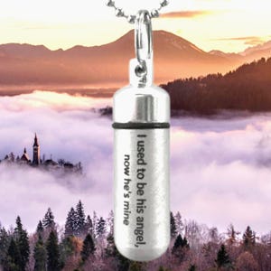 May include: A silver-colored, cylindrical pendant with a screw-top lid, suspended from a ball chain. The pendant has the engraved text: "I used to be his angel, now he's mine". The background features a scenic view of mountains and clouds.