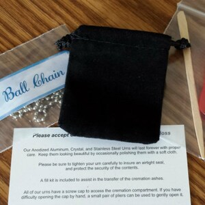 May include: A close-up of a 24-inch ball chain necklace, a black velvet pouch, a wooden stick, and a red plastic tool. The text "Please accept..." is visible on a white piece of paper.