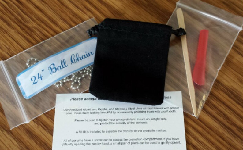 May include: A close-up of a 24-inch ball chain necklace, a black velvet pouch, a wooden stick, and a red plastic tool. The text "Please accept..." is visible on a white piece of paper.