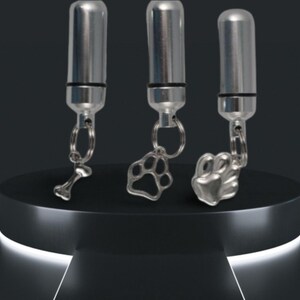 May include: Three silver pet urn keychains displayed on a black pedestal. Each keychain has a cylindrical urn attached to a ring, with charms shaped like a bone, a paw print, and a paw. The background is dark.