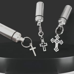 May include: Three silver-colored, cylindrical keychain pendants with cross charms. The pendants have a screw-top design and are attached to keyrings or chains. The crosses vary in style, with one featuring an openwork design.
