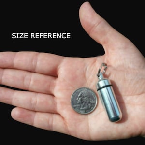 Pode incluir: Um pequeno recipiente cil&iacute;ndrico para comprimidos em metal prateado com um anel de veda&ccedil;&atilde;o de borracha preta e um anel de fixa&ccedil;&atilde;o met&aacute;lico no topo. O recipiente &eacute; mostrado ao lado de uma moeda como refer&ecirc;ncia de tamanho.