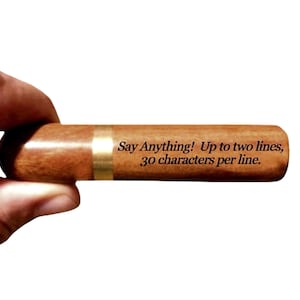 May include: A wooden cigar-shaped object with a brass band. The text "Say Anything! Up to two lines, 30 characters per line" is engraved on the surface. The item is held by a hand against a white background.