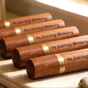 May include: Five cylindrical wooden keepsakes, each featuring a brass band and the inscription "In Loving Memory." The items are a rich brown hue and are displayed on a light-toned surface, possibly for remembrance.