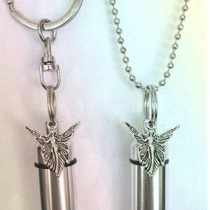 May include: Two silver-toned urn pendants. One hangs from a keychain, the other from a ball chain necklace. Each has a cylindrical body and a decorative fairy charm. The pendants are designed to hold a small amount of cremated remains.