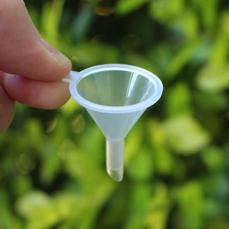 May include: A clear plastic funnel with a wide top and a narrow spout. The funnel is small and is being held by a hand.