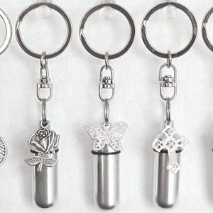 May include: Five silver-toned keychains with decorative pendants. The pendants include a tree of life, a rose, a butterfly, a cross, and a heart. Each keychain has a small, cylindrical container attached.