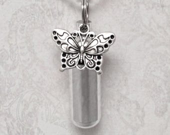May include: A silver-colored butterfly pendant with an intricate design, attached to a small, clear, cylindrical vial. The pendant is suspended from a silver chain, creating a delicate and ornamental piece. The vial is likely for holding small keepsakes.