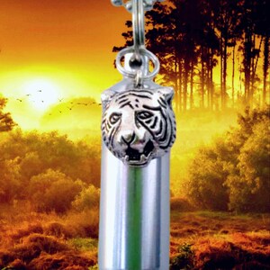 May include: Silver-colored pendant necklace featuring a tiger head design. The pendant is attached to a silver ball chain. The background is a sunset scene with trees and a golden sky.