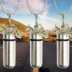 May include: Three silver necklaces with a motorcycle charm and a silver cylinder pendant. The necklaces are on a chain and are hanging against a blurred background of a road.