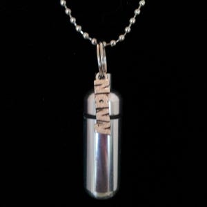 May include: A silver-colored cylindrical pendant with the word "NAVY" in capital letters, hanging from a ball chain necklace. The pendant has a screw-top design. The background is black.