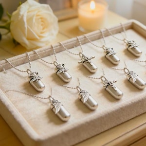 May include: A collection of silver-toned memorial necklaces displayed on a beige tray. Each necklace features a small, cylindrical pendant with a decorative floral or leaf design. A white rose and a lit candle are in the background.