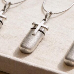 May include: Three silver-toned pendant necklaces with a cross design. Each necklace features a cylindrical pendant with a cross accent. The pendants are displayed on a light-colored surface, suggesting a jewelry display.