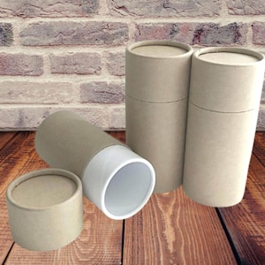 May include: A collection of cylindrical cardboard tubes and a small round container. The tubes are beige with matching lids, one is open, revealing a white interior. The items are arranged on a wooden surface against a brick wall.