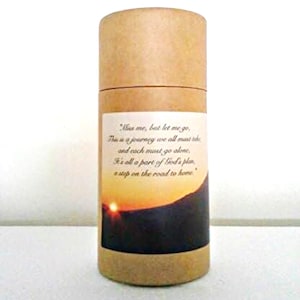May include: A cylindrical container made of tan cardboard. A label wraps around the container, featuring a sunset image and the text: "Miss me, but let me go, This is a journey we all must take and each must go alone, It's all a part of God's plan, a step on the road to home."