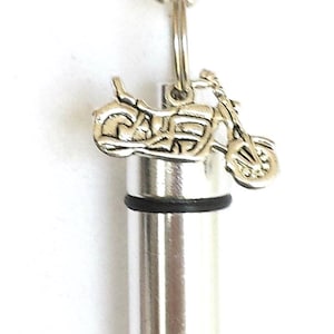 May include: Silver-toned motorcycle pendant necklace. The pendant features a miniature motorcycle charm above a cylindrical container. The necklace has a ball chain and a clasp. The background is white.