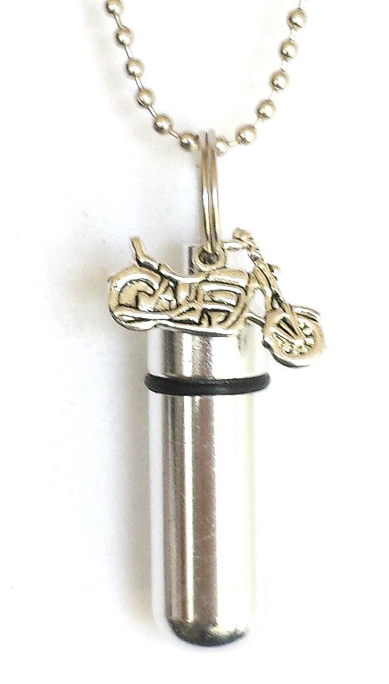 May include: Silver-toned motorcycle pendant necklace. The pendant features a miniature motorcycle charm above a cylindrical container. The necklace has a ball chain and a clasp. The background is white.