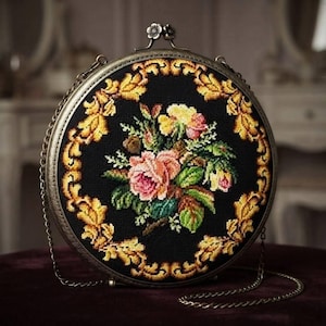 Puede incluir: Un bolso redondo de punto de cruz negro con un ramo central de rosas rosas y amarillas rodeado por un borde de hojas de color amarillo dorado. El bolso tiene un marco de metal color bronce, un cierre en forma de flor y una fina correa de cadena.
