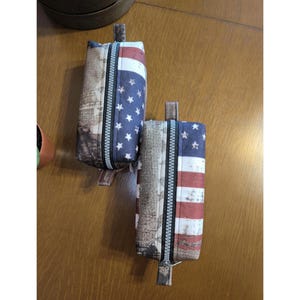 May include: Two rectangular pencil cases with zip closures. One features the American flag design with red and white stripes and blue stars. The other has a vintage map print.